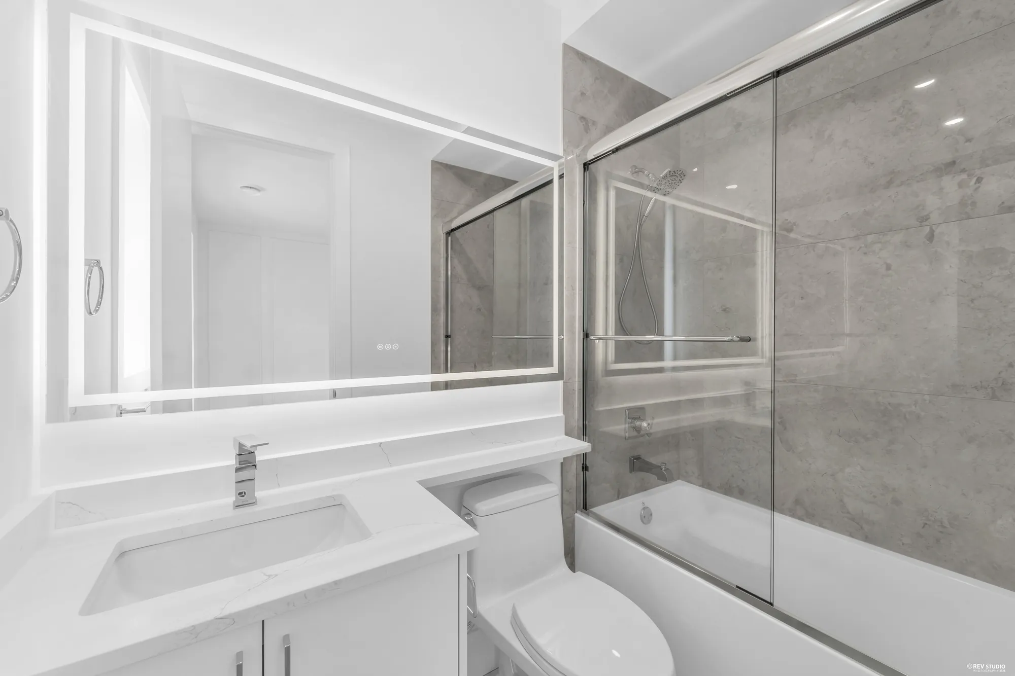 A bathroom with a tub and shower behind sliding glass, grey marble to the ceiling