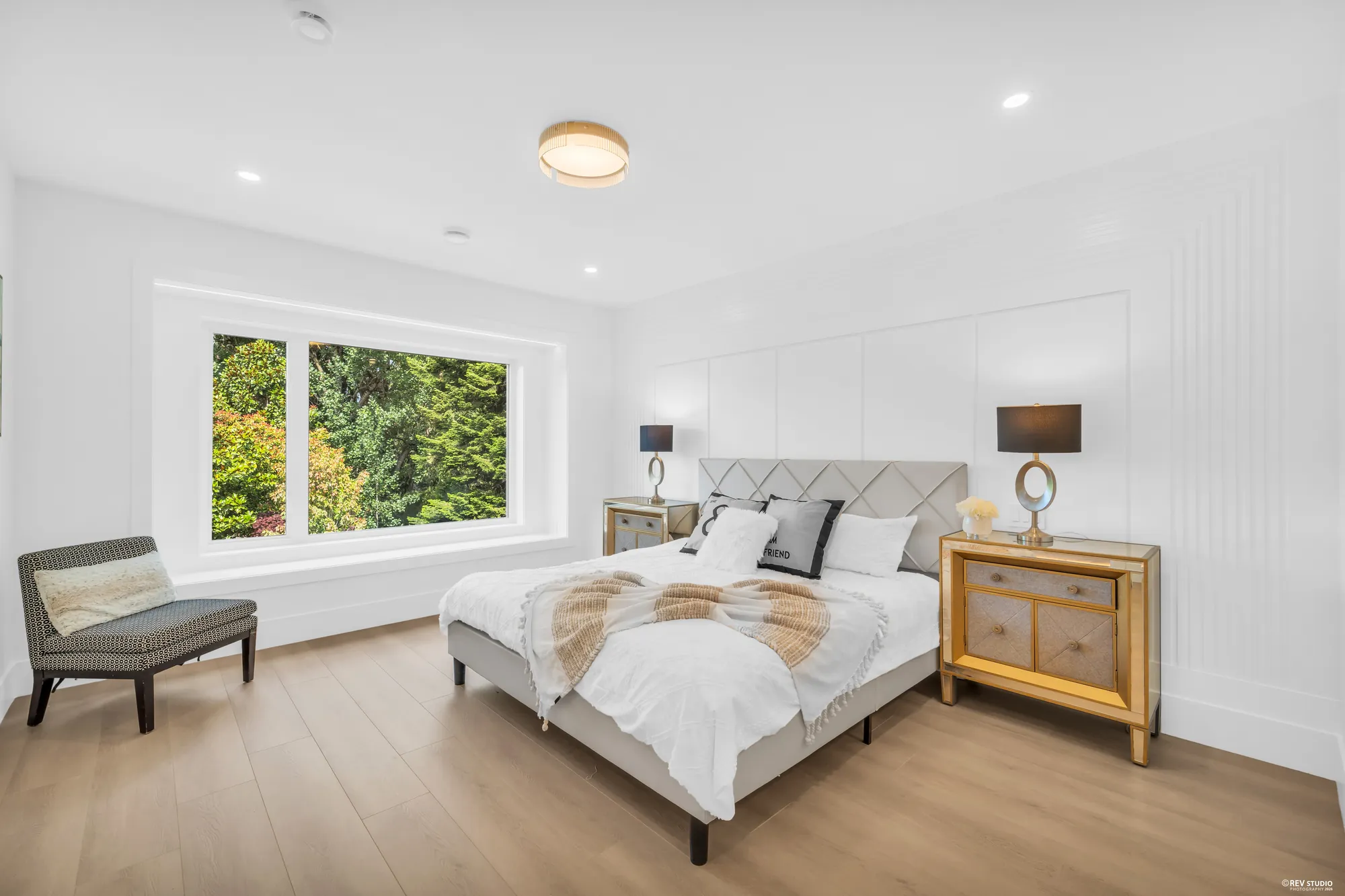 A dressed bedroom with a wide window onto the trees and a bench under it